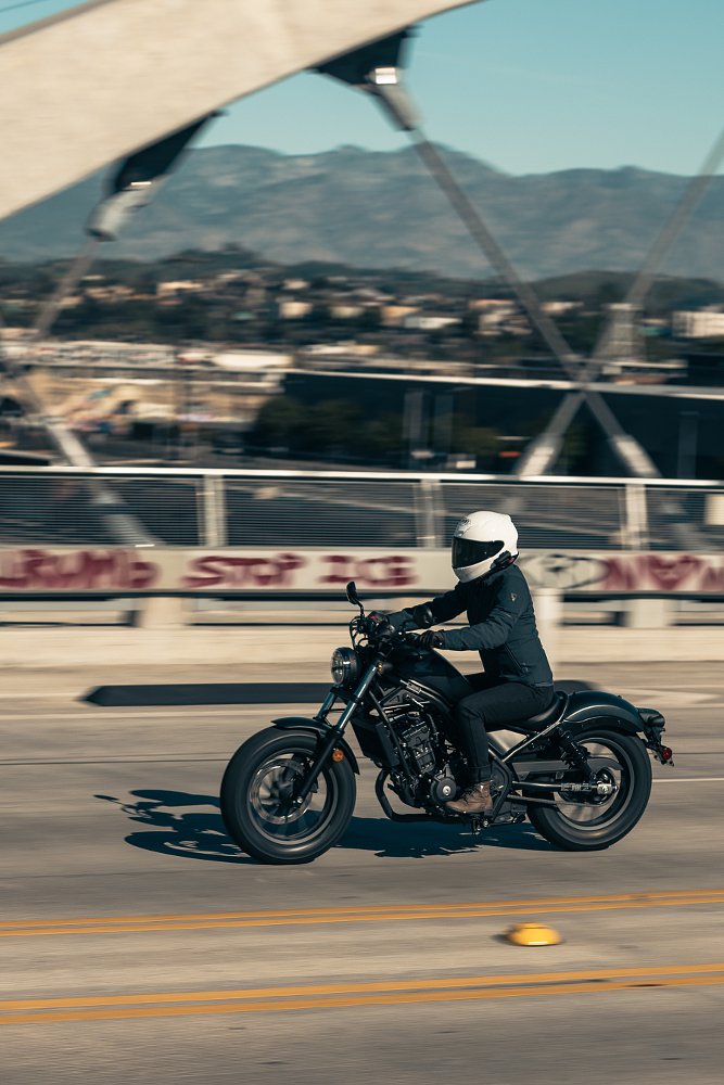 woman riding a black Rebel 300 across a city bridge